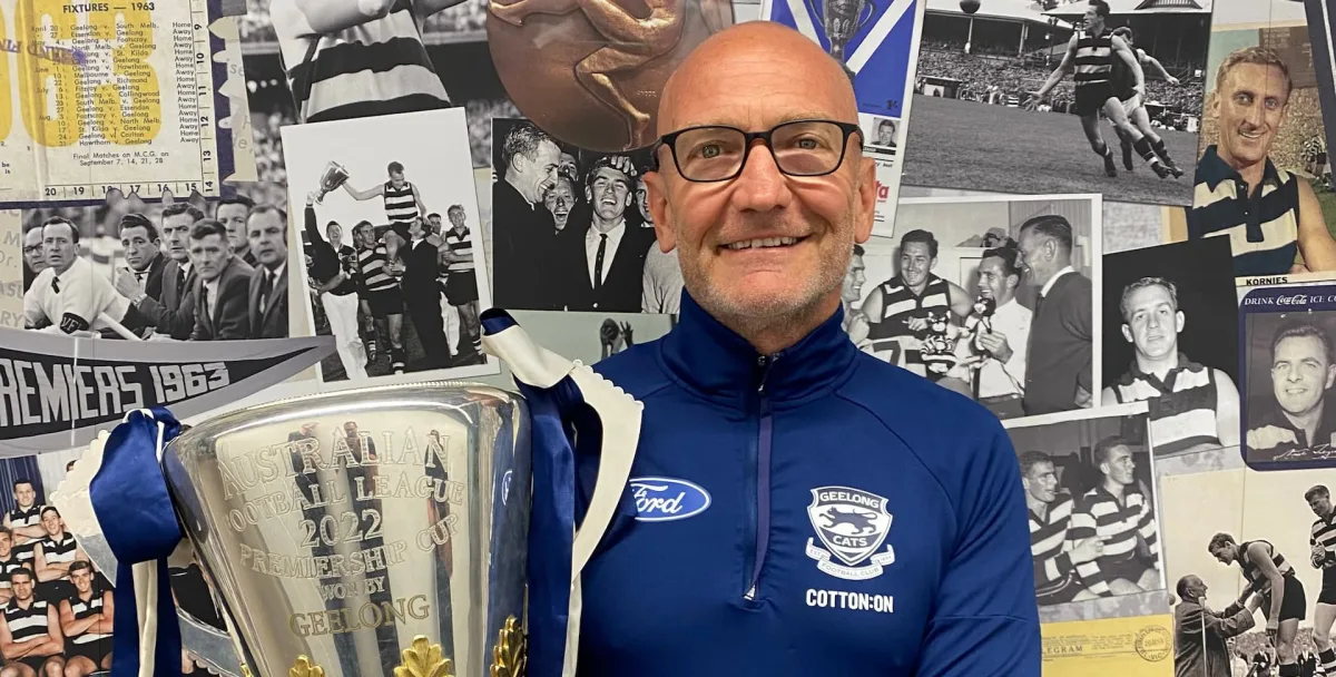 Dr Steve Saunders holding an AFL premiership cup in front of a Geelong Cats memorabilia wall